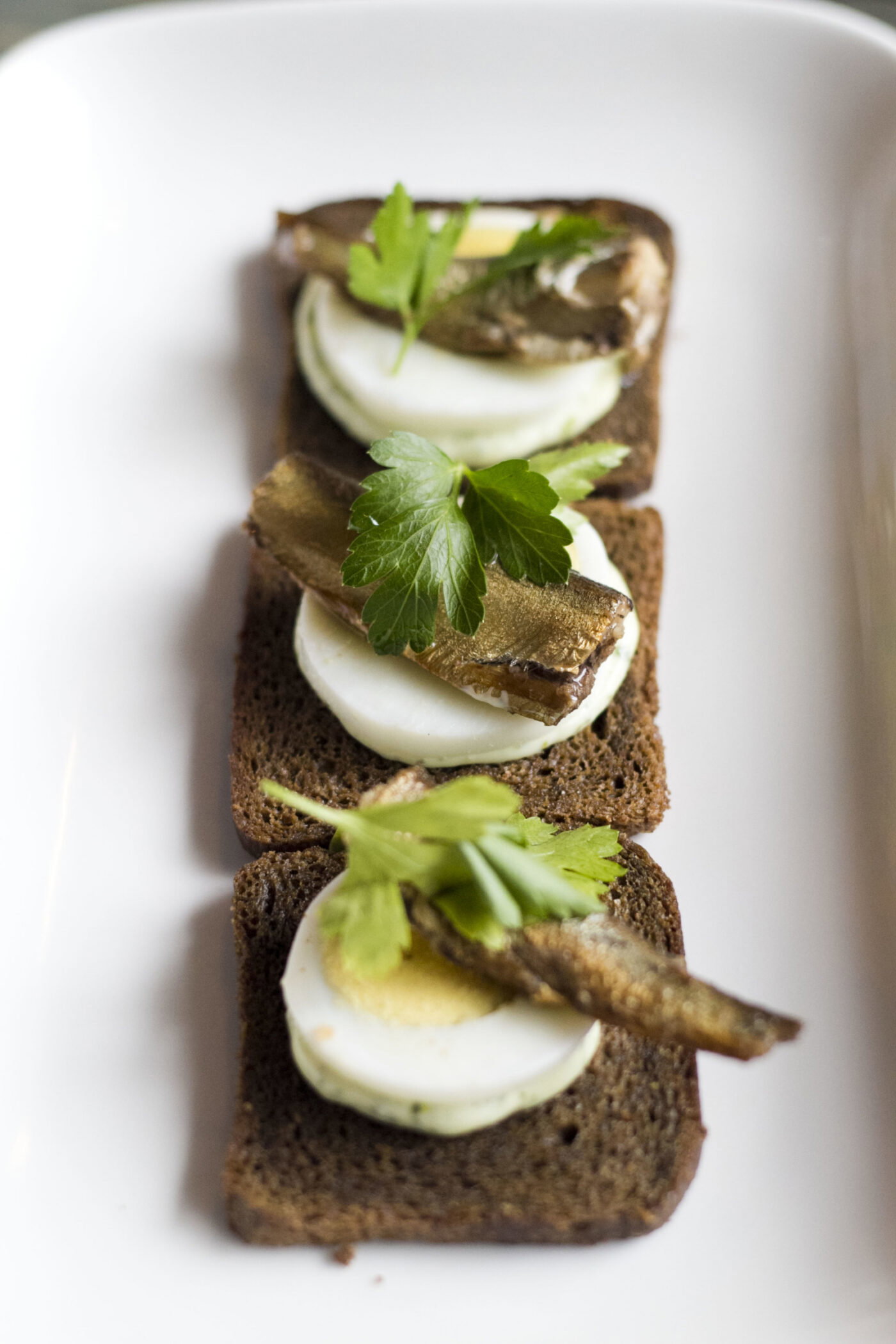 Baltic Sprat Buterbrodi: Tiny smoked fish, akin to mackerel, stacked on squares of pumpernickel toast holding eggs and a swath of parsley mayo.