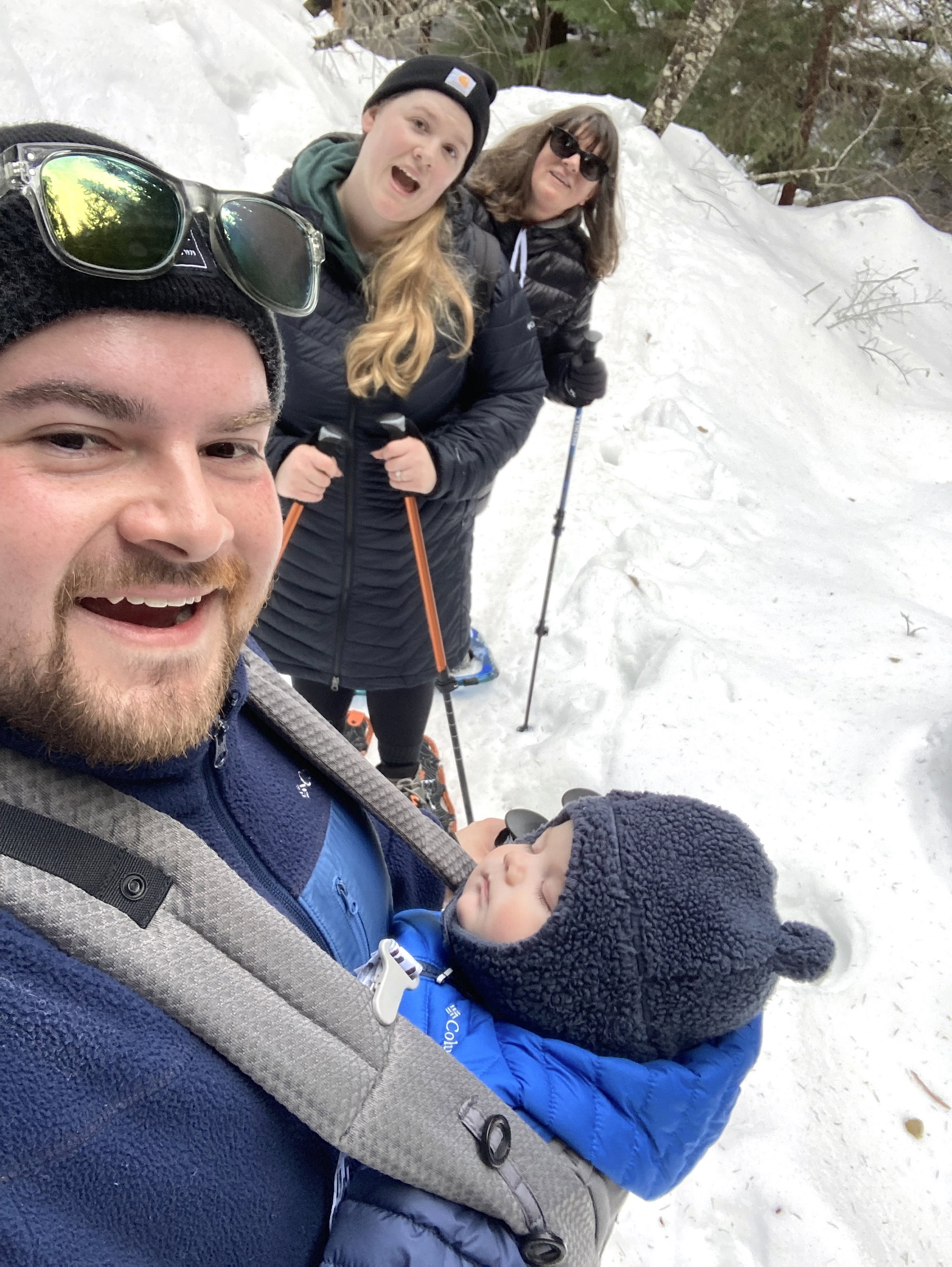 Man smiling with a sleeping baby in a chest carrier, with two women behind him holding poles and smiling, against a snowy backdrop